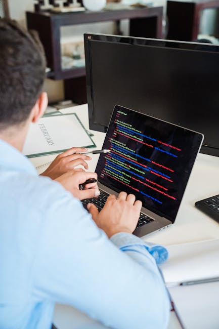 A desktop workspace featuring a laptop displaying code or text on its screen, placed on a dark wooden desk against a textured brick wall. To the right of the laptop, there is an open notebook with a b