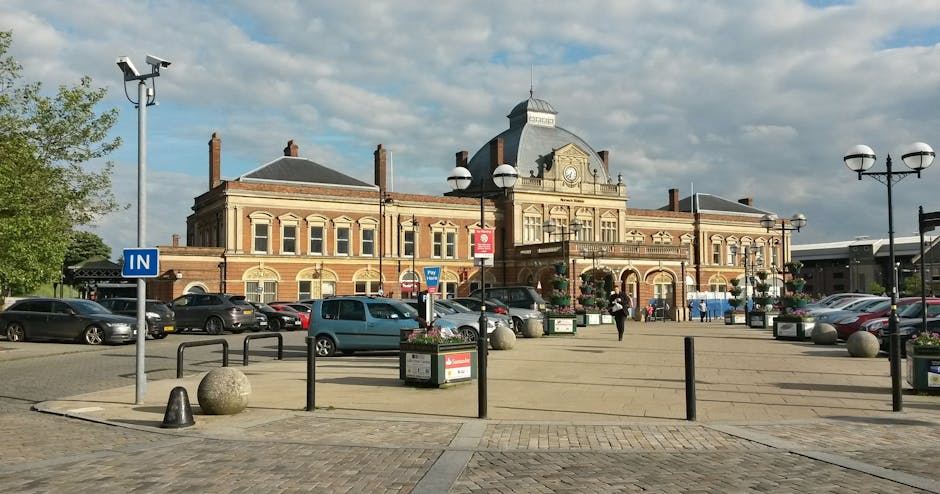 A large, historic brick building with ornate architectural details and a clock at the center, located on Blackstock Road near Finsbury Park station. In front of the building, there is a spacious paved area with parked cars along the perimeter. The scene includes several street lamps, planters with flowers, black metal posts, and bollards, with a few pedestrians walking. The sky is partly cloudy with patches of blue, indicating daylight, and the overall environment appears clean and well-maintained, reflecting a typical urban setting with attention to cleanliness and decor. This image conveys the exterior of a prominent local building in Finsbury Park, captured in natural lighting.