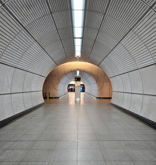 Interior of an empty subway train carriage with clean, patterned fabric seats arranged in rows along both sides of the vehicle, separated by a central aisle with a reflective, dark floor. The carriage features white plastic walls with blue handrails and vertical supports, large windows along each side, and bright overhead lighting. The surfaces appear well-maintained and free of dust or dirt. This image, associated with Finsbury Park Carpet Cleaners, highlights thorough surface cleaning and hygiene in public transportation settings, emphasizing the importance of sanitisation and deep cleaning for maintaining cleanliness standards.