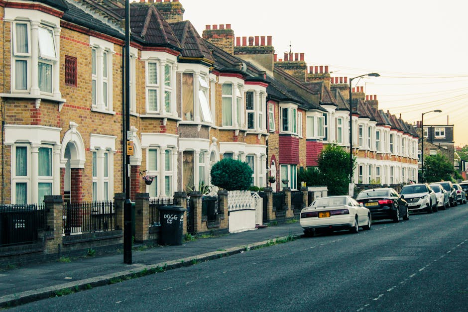 A row of Victorian terraced houses on Stroud Green Road in Finsbury Park, featuring brick facades with white bay window frames and decorative cornices. The houses have small front gardens enclosed by black iron fences, with some greenery such as bushes and small trees. The street is lined with parked cars along the curb, and the roadway is clean and well-maintained under soft natural lighting, typical of an early evening setting. The scene reflects a typical residential area in Finsbury Park with attention to surface cleanliness and structural maintenance, emblematic of the type of properties serviced by Finsbury Park Carpet Cleaners with professional surface cleaning and deep cleaning solutions.