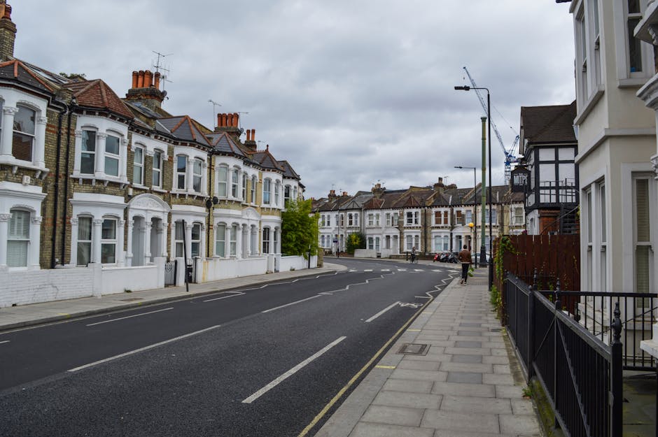 A quiet residential street on Stroud Green Road in Finsbury Park featuring Victorian terraces with ornate white facades, bay windows, and tiled roofs. The street is paved with asphalt and has marked parking bays along the curbside. Sidewalks are made of concrete slabs, with black metal railings and a few small trees lining the pavement. Overcast sky with grey clouds provides diffuse lighting, and there are no visible vehicles or pedestrians. The overall scene appears clean and well-maintained, reflecting typical urban residential surroundings. Finsbury Park Carpet Cleaners specializes in domestic cleaning and surface cleaning services in this area, ensuring hygiene and cleanliness for such Victorian property exteriors.